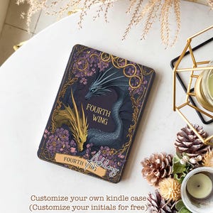 Puede incluir: Una funda Kindle personalizada con un fondo morado y un dise&ntilde;o de drag&oacute;n dorado y azul. El texto "FOURTH WING" est&aacute; impreso en la funda. El nombre "Ashley" est&aacute; escrito en cursiva en la esquina inferior derecha de la funda.