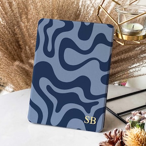 Puede incluir: Una funda para tableta con estampado azul y marino con las iniciales "SB" en dorado. La funda presenta un diseño ondulado y se muestra sobre una superficie blanca. La estética general es moderna y elegante.