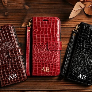 May include: Three rectangular wallet-style phone cases in dark brown, bright red, and black, each featuring a textured crocodile skin pattern, a gold-toned wrist strap, a magnetic closure tab, and the gold-embossed initials AB on the front cover.
