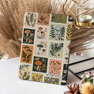 Puede incluir: Una funda rectangular con un collage de ilustraciones estilo sello postal de setas, helechos y flores en varios colores. El nombre "Ashley" está escrito en cursiva en la funda. La funda es probablemente para una tableta o un lector electrónico.