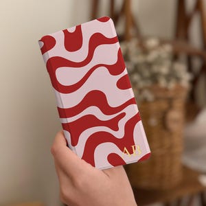 Pode incluir: Uma capa de telemóvel retangular com um padrão abstrato ondulado em vermelho escuro e rosa claro. O canto inferior direito apresenta as iniciais douradas AB numa fonte serifada. A capa tem um acabamento mate suave e um design fino.