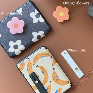 May include: Two phone cases with different designs. One case has a black background with white flowers and a pink flower phone grip. The other case has a white background with a banana pattern and a black phone grip. There are also two phone grips, one orange and one white.