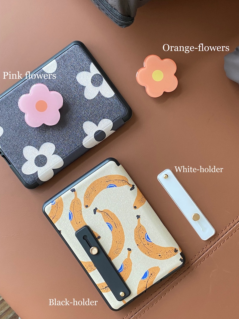 May include: Two phone cases with different designs. One case has a black background with white flowers and a pink flower phone grip. The other case has a white background with a banana pattern and a black phone grip. There are also two phone grips, one orange and one white.