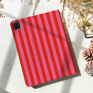 May include: A rectangular tablet case featuring a vertical striped pattern in alternating shades of bright red and pink. The case includes a square camera cutout in the upper left corner and the text Book back. printed in the upper left corner area.
