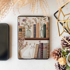 Pode incluir: Uma capa branca para Kindle com um design de estante de livros que apresenta um gato branco, flores e livros. A capa tem uma borda marrom e um fundo branco.