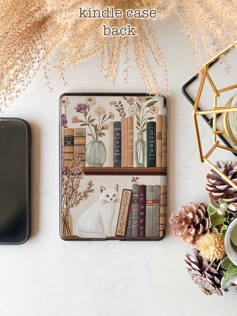 Pode incluir: Uma capa branca para Kindle com um design de estante de livros que apresenta um gato branco, flores e livros. A capa tem uma borda marrom e um fundo branco.