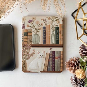 Pode incluir: Um gato branco est&aacute; sentado em uma prateleira com uma cole&ccedil;&atilde;o de livros e flores secas. A prateleira faz parte de uma capa para Kindle com um design floral.