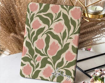 Peut inclure: Une housse de tablette à motif floral avec des fleurs roses et des feuilles vertes sur fond beige. Le nom "Ashley" est écrit en cursive. L'étui est rectangulaire avec un bord noir. Le design rappelle une impression botanique.
