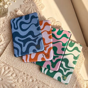 May include: Four rectangular e-reader cases featuring wavy, abstract patterns in various colour combinations. The cases display the initials JD, YM, TA, and CB in gold lettering. The colour schemes include blue, orange and lavender, pink and green, and green and white.