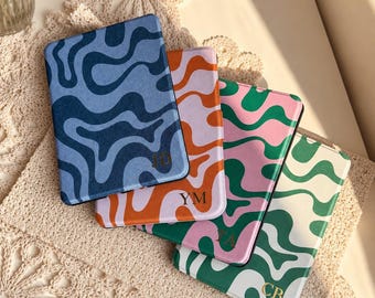 Puede incluir: Cuatro fundas rectangulares para lector electrónico con patrones abstractos ondulados en varias combinaciones de colores. Las fundas muestran las iniciales JD, YM, TA y CB en letras doradas. Los esquemas de color incluyen azul, naranja y lavanda, rosa y verde, y verde y blanco.