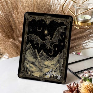 Peut inclure: Étui de tablette rectangulaire noir avec un motif de dragon et de montagnes dorées. Le dragon vole au-dessus des montagnes sous un ciel nocturne avec une lune et des étoiles. Le nom "Ashley" est écrit en cursive.