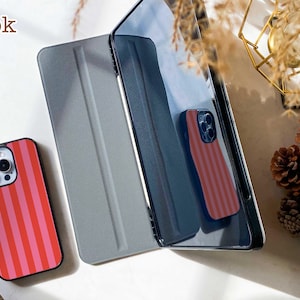 May include: A smartphone case with vertical red and pink stripes next to a dark gray tablet folio cover. The word book is printed in the upper left corner. The tablet cover has a textured surface and is positioned upright next to the phone case.