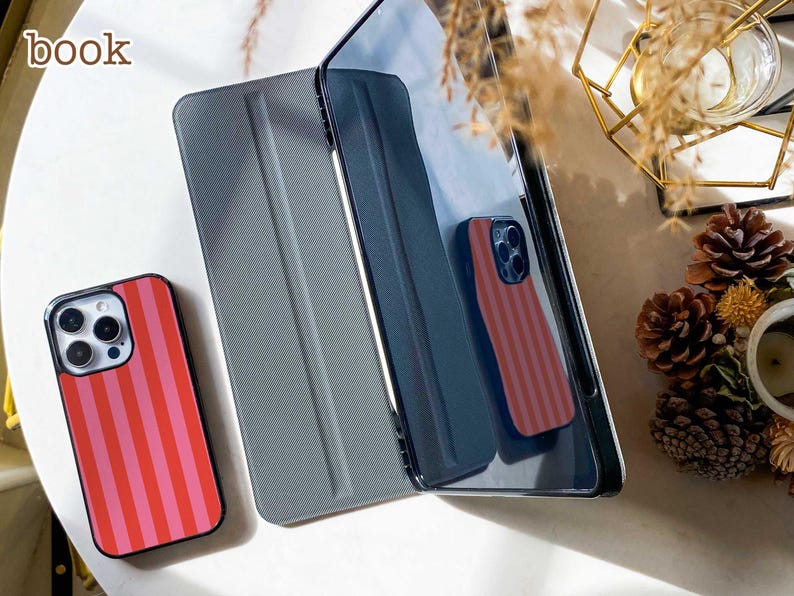 May include: A smartphone case with vertical red and pink stripes next to a dark gray tablet folio cover. The word book is printed in the upper left corner. The tablet cover has a textured surface and is positioned upright next to the phone case.