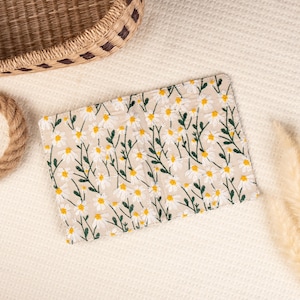 May include: A small, beige fabric wallet with a white daisy floral pattern. The wallet is closed and lying on a white, textured surface.