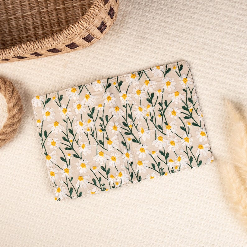 May include: A small, beige fabric wallet with a white daisy floral pattern. The wallet is closed and lying on a white, textured surface.