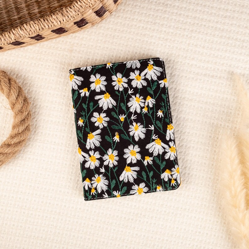 May include: A black passport holder with a white daisy floral embroidery pattern.