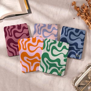 May include: Five square e-reader cases featuring wavy, abstract patterns in various colour combinations including maroon, light purple, blue, orange, and green. Two of the cases display gold-coloured initials, specifically SB and SA.