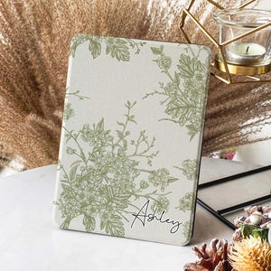 Peut inclure: Une housse pour liseuse à motifs floraux blancs et verts avec le nom "Ashley" imprimé dans le coin inférieur droit.