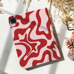 May include: A tablet case featuring a wavy, abstract pattern in bold red and light pink. The case has a rectangular shape with a dedicated cutout for a camera lens in the top left corner. The text "Book back." is printed in the top left corner.