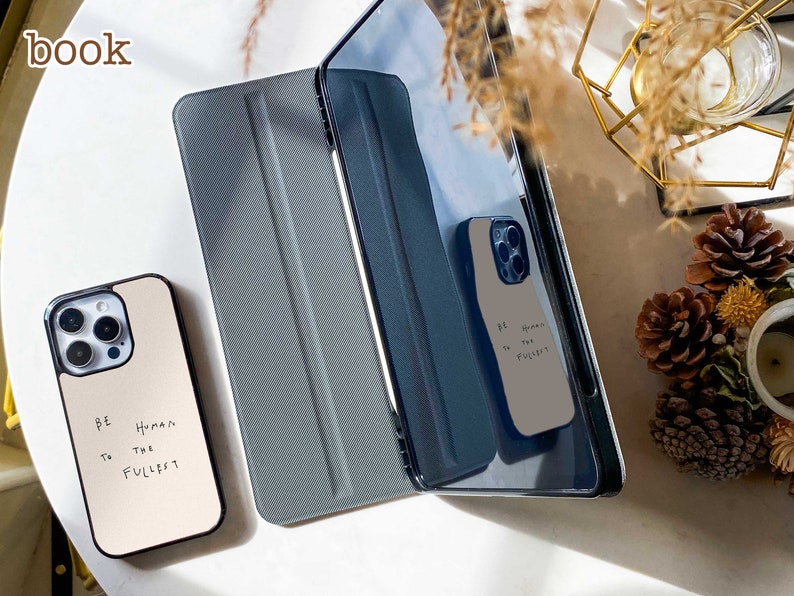 May include: Two phone cases with a white background and black text that reads "Be Human to the fullest". The cases are on a white surface with a gray and black tablet case.