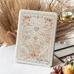 May include: An off-white rectangular case with a floral and ribbon design. The case has a lace border and a pattern of roses, doves, and bows in soft pink and green. The name "Ashley" is written in cursive on the front.