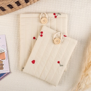 以下が含まれることがあります： チェリー刺繍と木製ボタン留めが施された、ベージュのキルティングポーチ2つ。ポーチは柔らかく質感のある生地で作られており、控えめなヴィンテージな雰囲気があります。