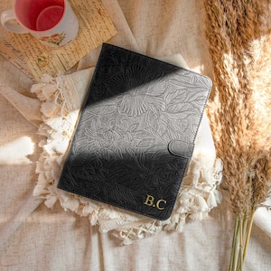 Puede incluir: Funda para tablet de cuero negro con un diseño en relieve floral y las iniciales doradas "B.C." en la parte delantera.