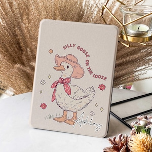 Puede incluir: Una funda beige para Kindle con una ilustración de dibujos animados de un ganso con un sombrero rosa y una bufanda roja. El texto "Silly Goose on the Loose" está escrito en un círculo alrededor del ganso. El nombre "Ashley" está escrito en cursiva en la parte inferior de la funda.