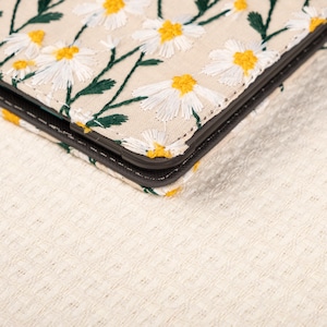 May include: A close-up of a beige fabric case with a floral pattern. The case is embroidered with white daisies and green stems. The case has a black border and is folded in half.