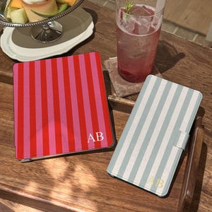 May include: A square tablet case with vertical red and pink stripes featuring the white monogram AB, and a rectangular phone case with vertical light blue and white stripes featuring the gold monogram AB, both with a smooth, matte finish.