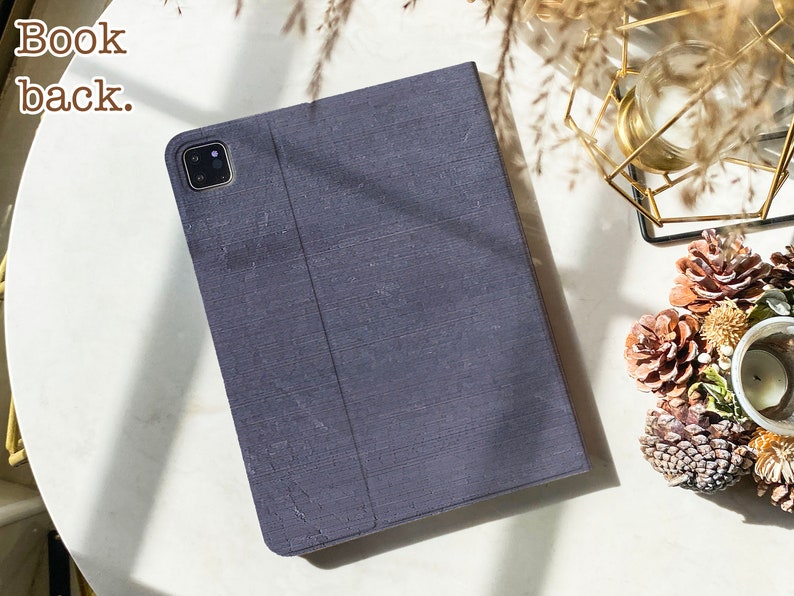 May include: A dark gray fabric iPad case with a camera lens cutout. The case is closed and the back is visible. The text "Book back." is visible in the top left corner of the image.