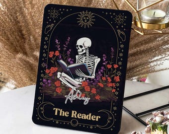 Puede incluir: Una carta de tarot negra y dorada con un esqueleto leyendo un libro. La carta tiene un diseño floral y el texto "Ashley The Reader".