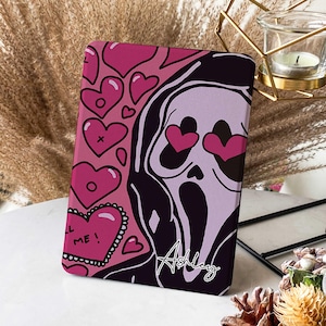 May include: A pink and black tablet case with a graphic of a ghost wearing a black robe and heart-shaped eyes. The ghost is surrounded by pink hearts and the text "Kill Me!" is on a pink heart in the lower left corner. The name "Ashley" is written in black cursive on the bottom right corner.