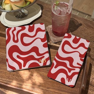 May include: A square tablet case and a rectangular phone case, both featuring a wavy red and light pink pattern. Each item is personalized with the gold-colored initials AB in the bottom corner. The cases have a smooth, matte finish.