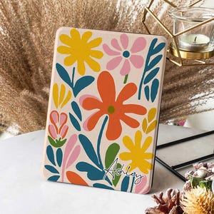 May include: A personalized Kindle cover with a floral design featuring pink, yellow, orange, green, and blue flowers on a cream background. The name "Ashley" is written in cursive at the bottom of the cover.