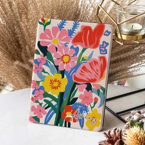 Può includere: Una custodia personalizzata per e-reader con un design floreale. La custodia ha uno sfondo bianco con un bouquet di fiori colorati in rosa, rosso, giallo e blu. Il nome "Ashley" è scritto in corsivo nero nella parte inferiore della custodia.