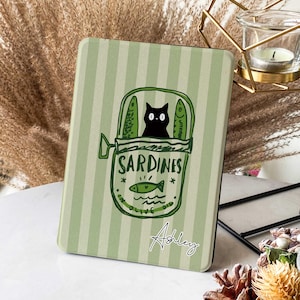 Puede incluir: Una funda a rayas verdes y blancas con un diseño de lata de sardinas de dibujos animados. Un gato negro es visible dentro de la lata, que lleva la etiqueta "Sardinas en aceite de oliva". El nombre "Ashley" está escrito en cursiva.