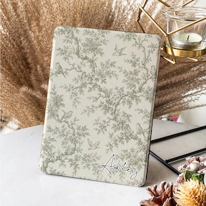 Könnte beinhalten: Eine Tablet-Hülle mit einem Toile-de-Jouy-Muster mit Vögeln und Blättern in Grau- und Weißtönen. Der Name "Ashley" ist in Schwarz in der unteren rechten Ecke geschrieben. Die Hülle wird auf einer weißen Oberfläche präsentiert.