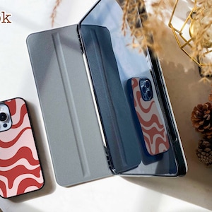 May include: A smartphone case featuring a wavy red and light pink pattern, positioned next to a dark grey tablet cover. The word book is printed in the upper left corner. The smartphone case is also reflected in the dark screen of the tablet.