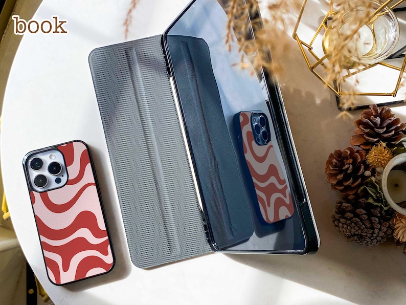 May include: A smartphone case featuring a wavy red and light pink pattern, positioned next to a dark grey tablet cover. The word book is printed in the upper left corner. The smartphone case is also reflected in the dark screen of the tablet.