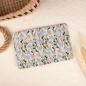 May include: A small, rectangular, beige fabric pouch with a blue and green floral embroidery pattern. The pouch is lying flat on a white surface.