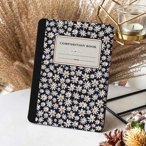May include: A black composition book with a white daisy pattern. The book has a label that says "Composition Book" and a space for a name. The name "Adley" is written in the space.
