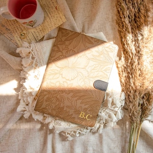 Puede incluir: Un diario de cuero marrón con un diseño floral en relieve y las iniciales "B.C." en letras doradas. El diario está sobre una superficie de tela beige.