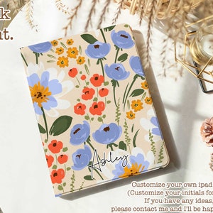 Op de afbeelding: Een gepersonaliseerde iPad-hoes met een bloemenpatroon met blauwe, rode, gele en witte bloemen op een crèmekleurige achtergrond. De naam "Ashley" staat in cursief op de hoes. Personaliseer je eigen iPad-hoes met je initialen gratis.