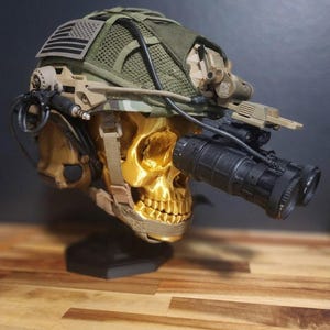 Pu&ograve; includere: Un teschio color oro con un elmetto in stile militare con visori notturni. L'elmetto &egrave; verde oliva con una parte superiore in rete e una toppa con la bandiera americana. Il teschio &egrave; montato su un supporto nero.