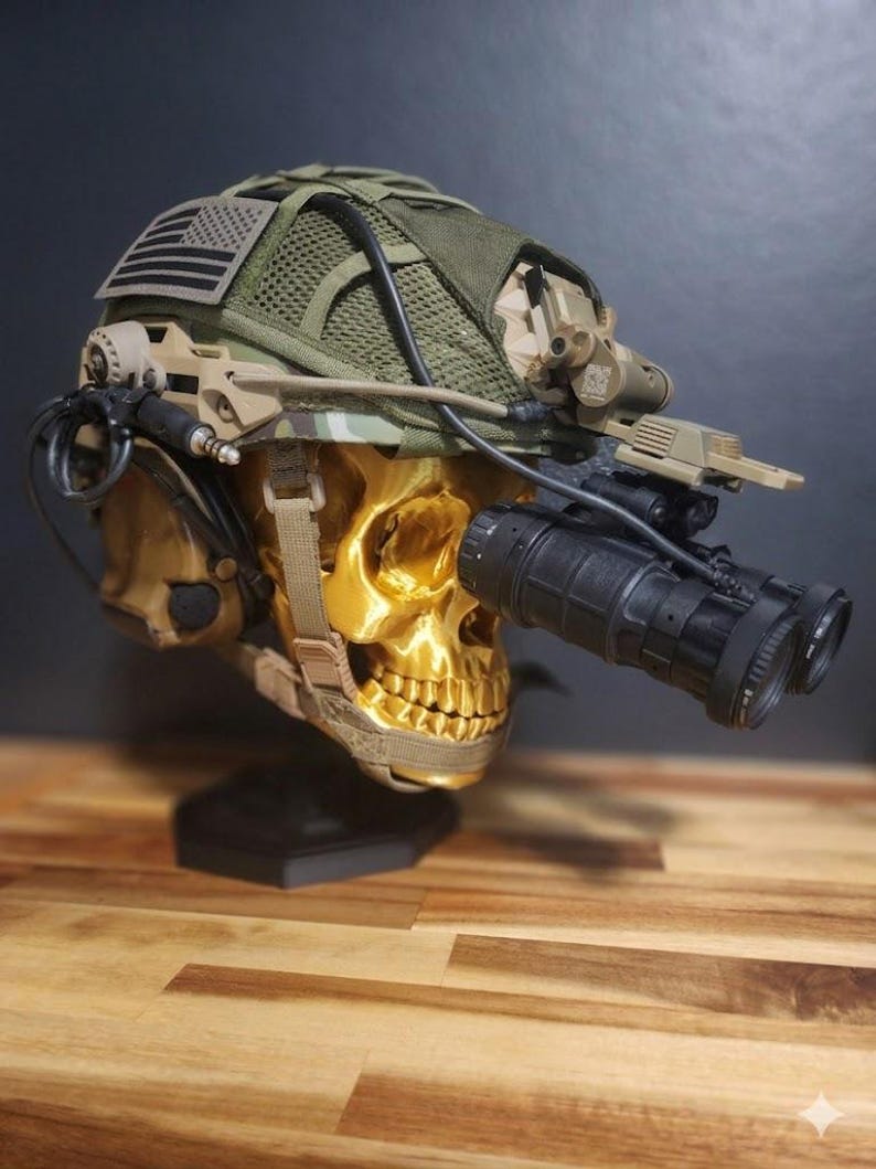 Pu&ograve; includere: Un teschio color oro con un elmetto in stile militare con visori notturni. L'elmetto &egrave; verde oliva con una parte superiore in rete e una toppa con la bandiera americana. Il teschio &egrave; montato su un supporto nero.