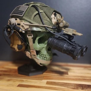 Pu&ograve; includere: Un casco verde a forma di teschio con dettagli mimetici, dotato di visori notturni, una toppa con la bandiera americana e vari accessori tattici. Il casco &egrave; esposto su un supporto nero, su uno sfondo scuro.