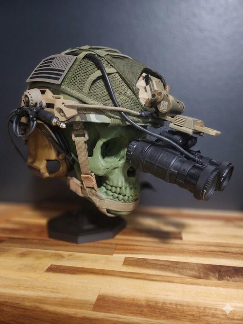 Pu&ograve; includere: Un casco verde a forma di teschio con dettagli mimetici, dotato di visori notturni, una toppa con la bandiera americana e vari accessori tattici. Il casco &egrave; esposto su un supporto nero, su uno sfondo scuro.