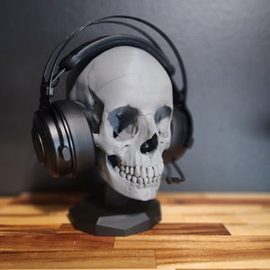 Pu&ograve; includere: Un supporto per cuffie a forma di teschio grigio con una cuffia nera. Il supporto ha una base esagonale. La cuffia ha grandi padiglioni auricolari e un microfono. Lo sfondo &egrave; scuro. Il supporto &egrave; su una superficie di legno.