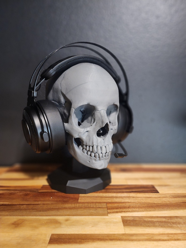 Pu&ograve; includere: Un supporto per cuffie a forma di teschio grigio con una cuffia nera. Il supporto ha una base esagonale. La cuffia ha grandi padiglioni auricolari e un microfono. Lo sfondo &egrave; scuro. Il supporto &egrave; su una superficie di legno.